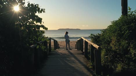 woman stands on wooden deck by the beach at sunrise golden hour using smartphone with running suit and sneakers