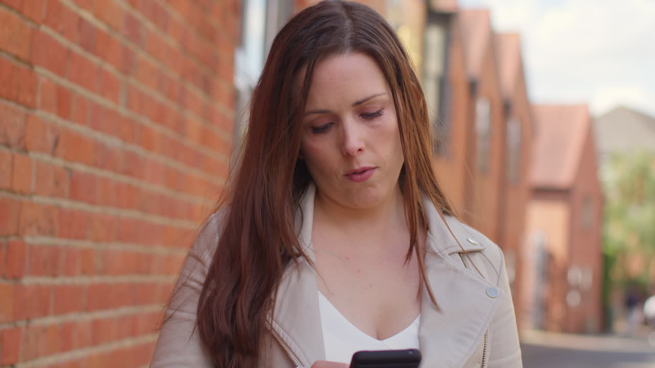 mujer al aire libre caminando a trabajar a lo largo de la calle de la ciudad alineada con casas mirando el teléfono móvil 2