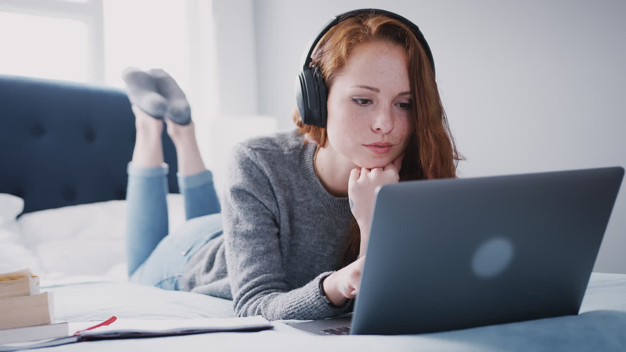 Female College Student Wearing Headphones Works On Bed In Shared House With Laptop And Mobile Phone