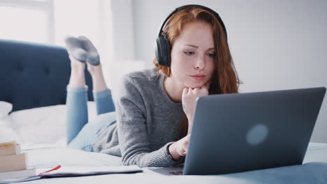 estudiante universitaria que usa auriculares trabaja en la cama en una casa compartida con portátil y teléfono móvil