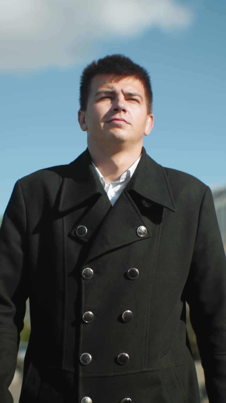 검은 코트를 입은 남자가 은 파란 하늘 아래 현대적인 사무실 건물, 움직이는 자동차, 그리고 활기찬 나무가 있는 바쁜 도시 도로를 따라 자신감 있게 고 있습니다.