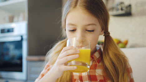 Close-up-of-the-nice-blonde-schoolgirl-drinking-orange-juice-from-the-glass-in-the-kitchen-in-front-of-the-camera.-Portrait-shot.-Indoors