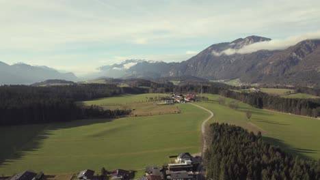 Luftdrohne-Der-Wunderschönen-Alpinen-Bergkette-In-Der-Ferne-Mit-Malerischen-Grasfeldern,-Bäumen-Und-Einer-Stadt