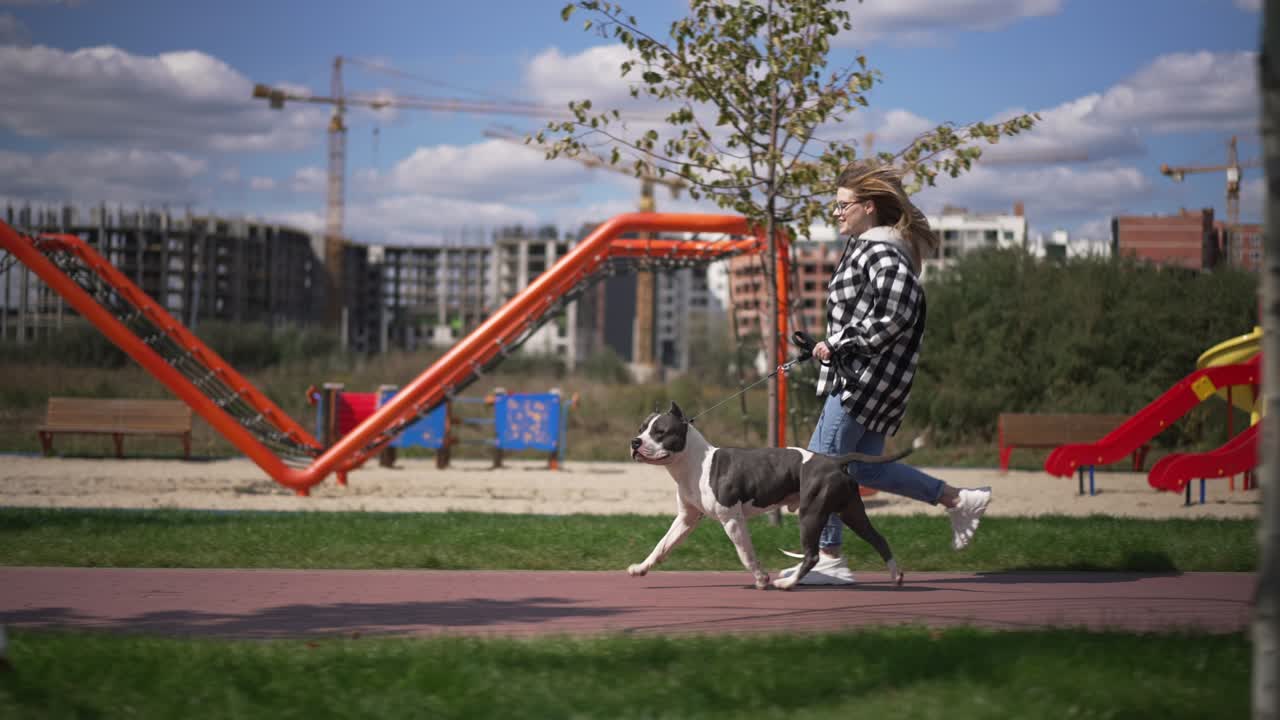 disparo ancho feliz emocionada mujer joven corriendo con el perro en un día soleado al aire libre pasando bancos en el frente. vista lateral seguimiento disparo de propietario caucásico positivo y mascota corriendo en cámara lenta en el sol.