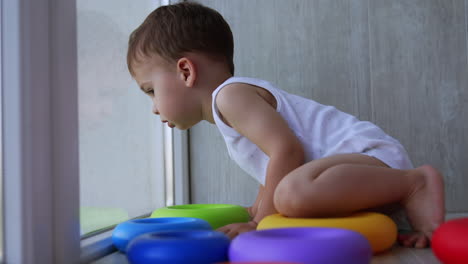 cute caucasian child sits on the floor near panoramic window. kid looks down first and then goes back to his toys.