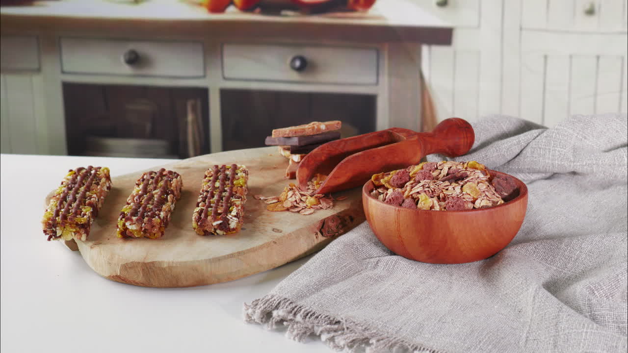 Camera moves closer to reveal granola bars, stacked chocolate, a wooden scoop, and a cereal bowl on a rustic wooden board, with soft kitchen textures and muted tones enhancing the cozy setting.