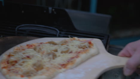 man putting a homemade pizza into a wood fired pizza grill oven on a wooden plate