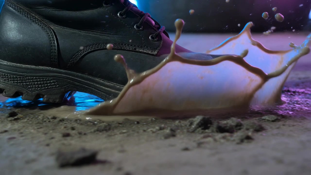 Sepatu bot hitam melayang menekan genangan lumpur saat melintas di studio, memancarkan cahaya biru magenta.