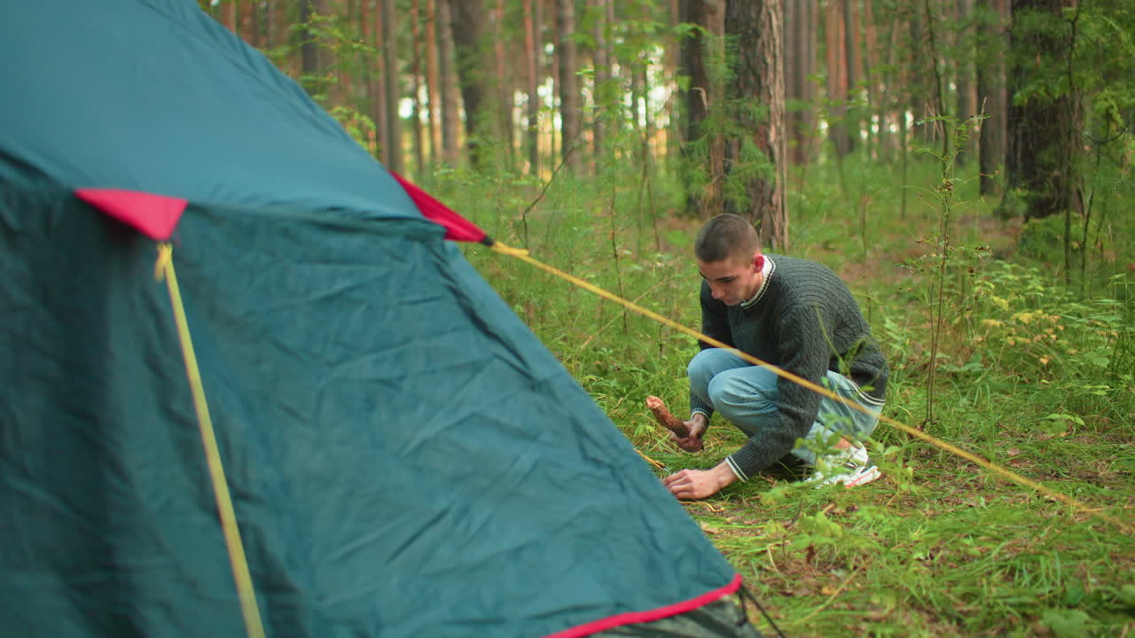Man in green sweater and jeans squats beside tent in forest while hammering peg into ground with wooden stick, securing yellow rope to stabilize camping setup among tall trees