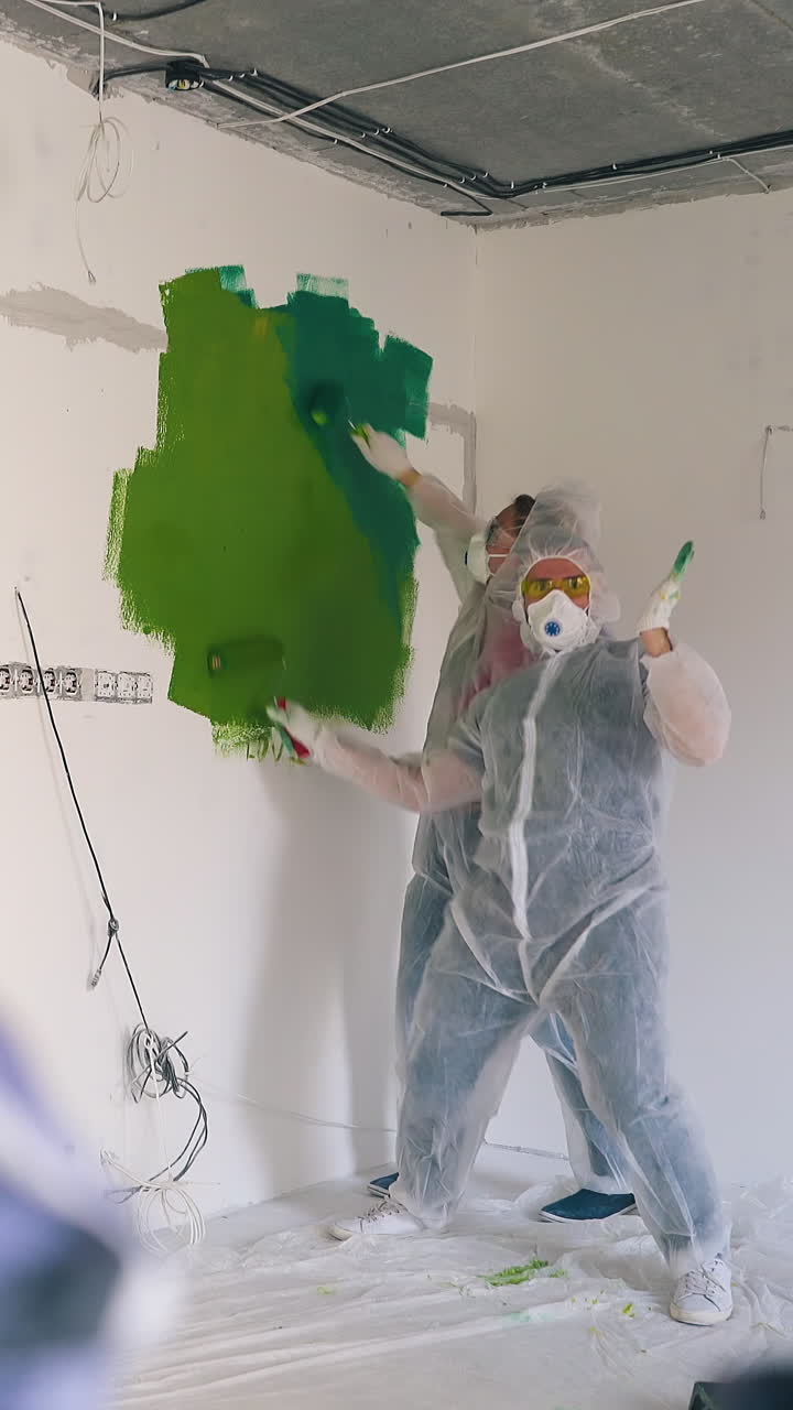 Dolly cam: joyful couple in transparent coveralls and respirators runs waving hands to paint white wall with rollers in room