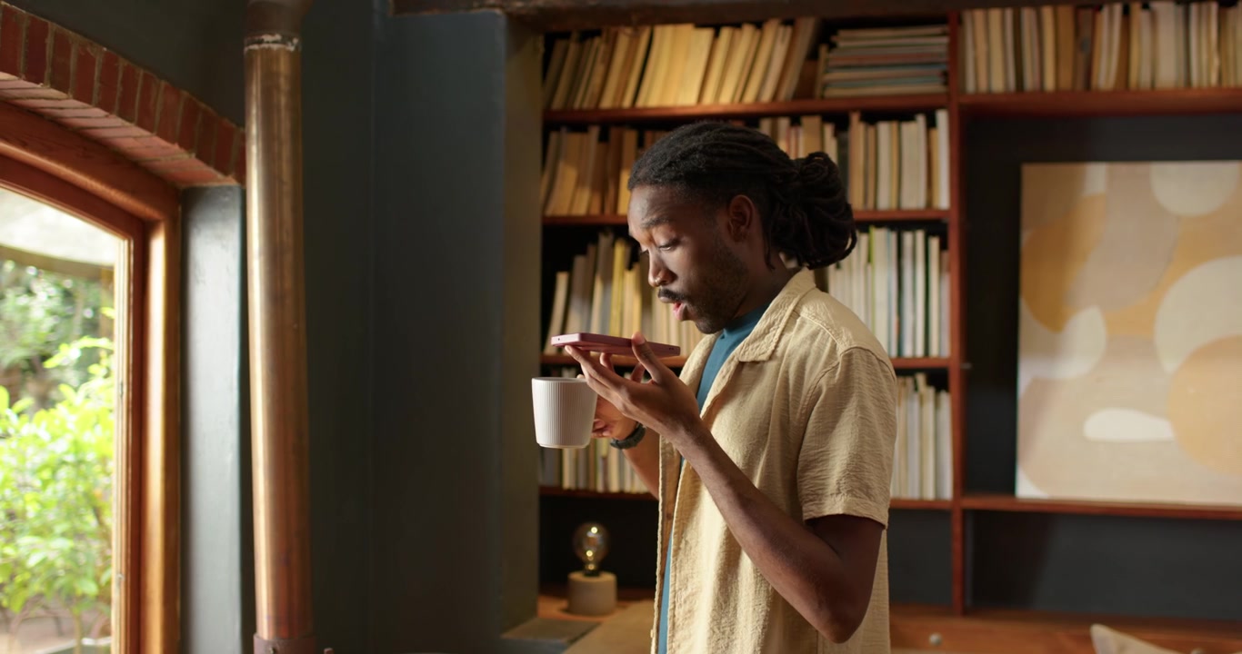 Homme en chemise beige regardant par la fenêtre, tenant une tasse sur une soucoupe, levant la tasse pour sentir la boisson.