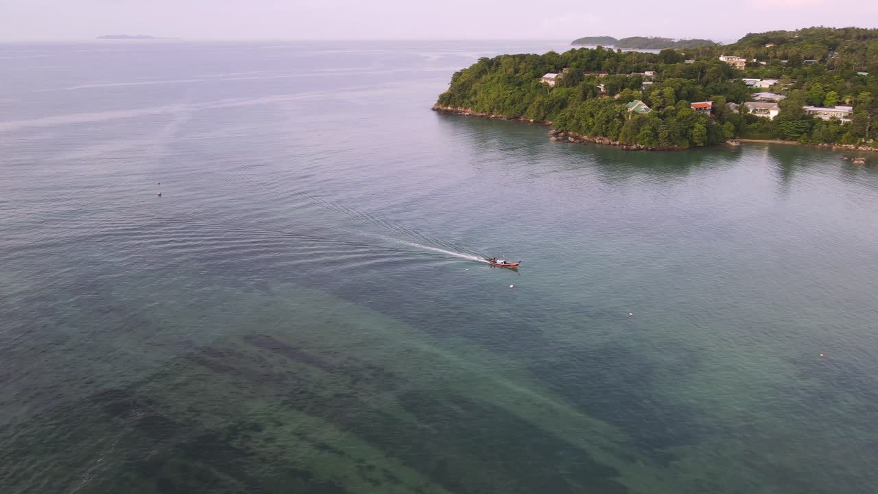 Ripresa aerea con drone sopra una spiaggia della Thailandia, oceano blu tropicale con una barca che naviga e una montagna verdeggiante.