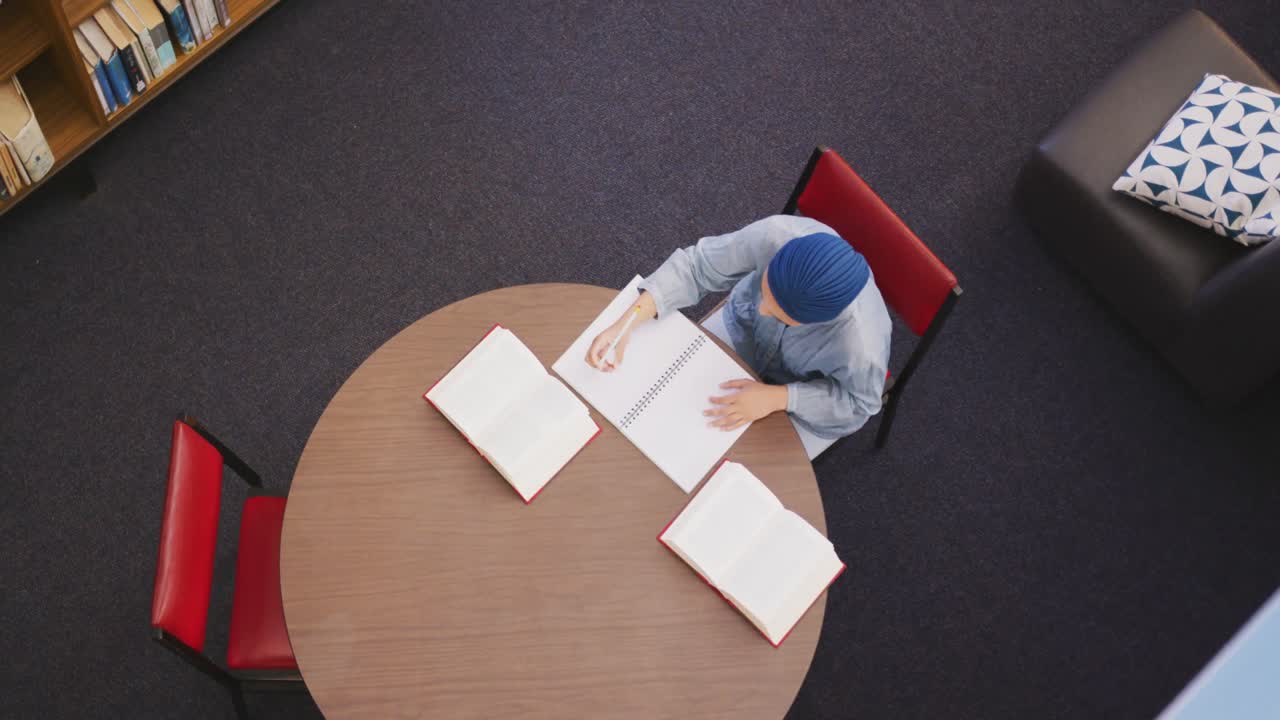 Asian female student wearing a blue hijab sitting at a round table and reading a book