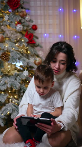 brunette woman in white sweater holds a cute toddler boy. mom and kid use a smartphone sitting at christmas tree. vertical video.