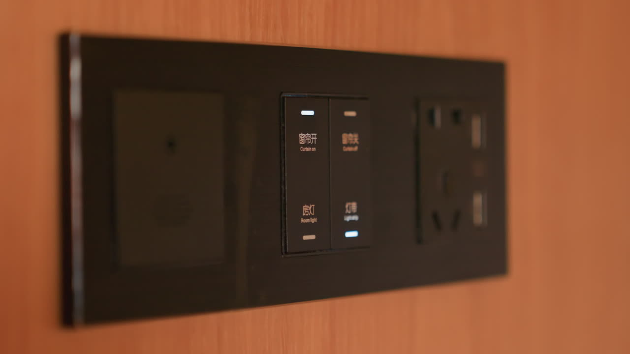 Hotel room control panel closeup, black power panel mounted on wood wall. Hand with ring touches illuminated touch buttons, USB ports and universal outlet visible. Warm ambient lighting creates cozy