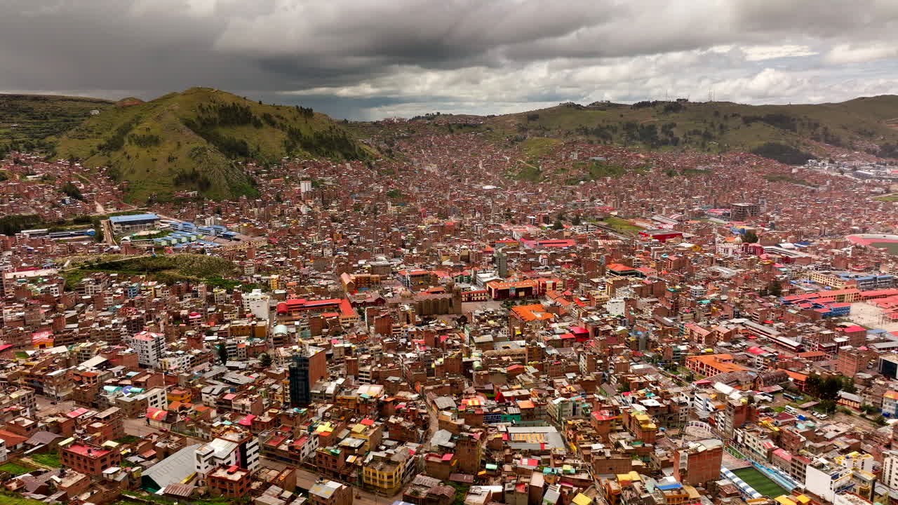 Panoramic aerial shot of Puno city in southeastern Peru. Dense brick buildings and red roofs cover the hillsides near Lake Titicaca under a cloudy sky. A vast high-altitude urban landscape