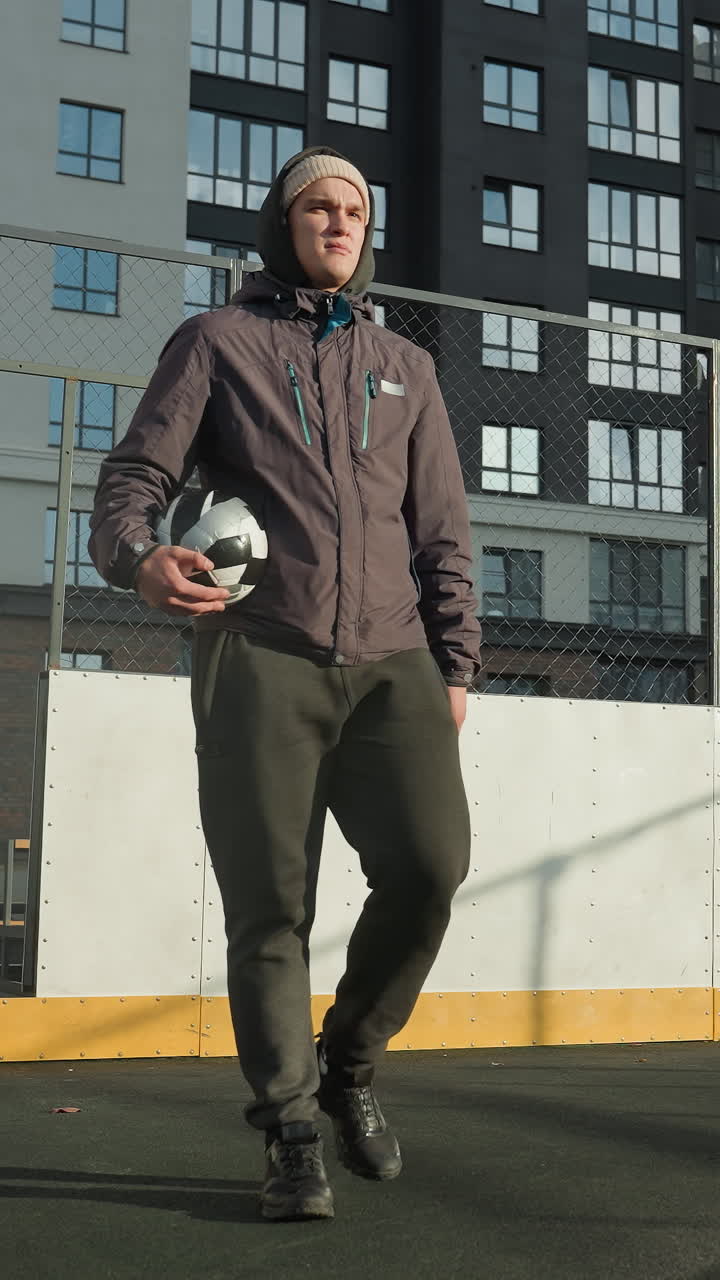 Footballer in hoodie and beanie holding ball in right hand walks along football pitch with goal post, tall residential building in background, and sunlight casting shadow on ground