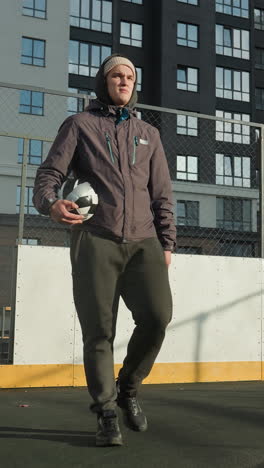 futbolista con capucha y gorra sosteniendo la pelota en la mano derecha camina a lo largo del campo de fútbol con poste de gol, edificio residencial alto en el fondo, y la luz del sol proyectando sombra en el suelo