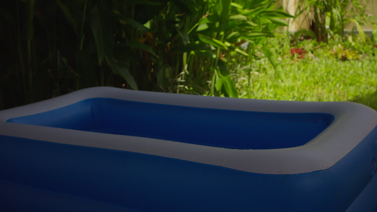 Ampia vista di una piscina gonfiabile blu posizionata in un giardino tropicale soleggiato con piante verdi ed erba, creando un'atmosfera serena per il tempo libero all'aperto.