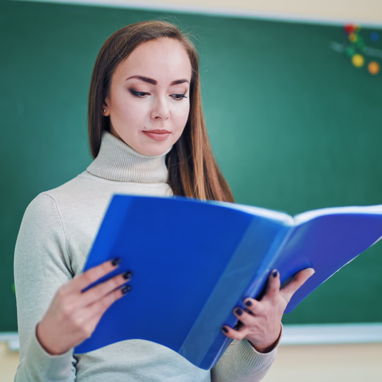 Beautiful teacher with a folder. Pretty young woman holding a blue folder and looking at files on the background of a board. Education concept.