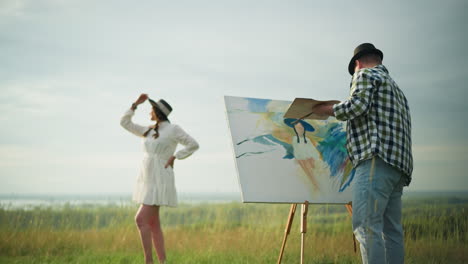 a painter in a checkered shirt and jeans creates a colorful portrait of a woman posing in a white dress and hat in a lush grassy field. the serene outdoor setting enhances the artistic moment