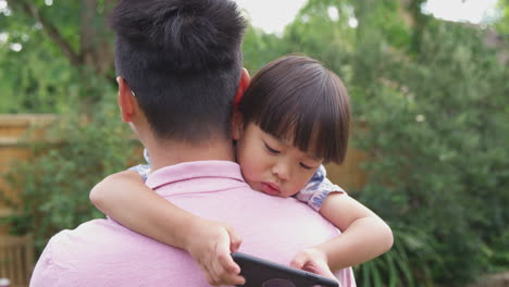 asian father cuddling son in garden as boy looks over his shoulder at mobile phone