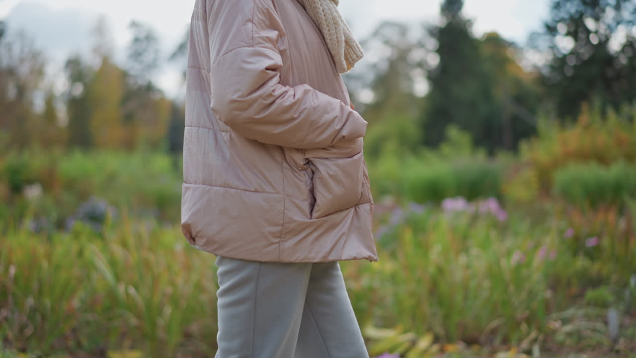close up of young lady walking thoughtfully along flower strewn garden path wearing hoodie and neck muffler with hand in hood pocket, grey jogger pants and sneakers, slight blur background