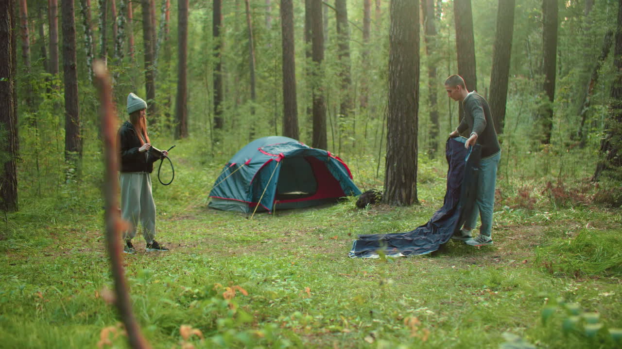 Woman in knit beanie watches man unfold tent fabric on grassy forest clearing with pitched tent in background surrounded by trees as they prepare gear and equipment