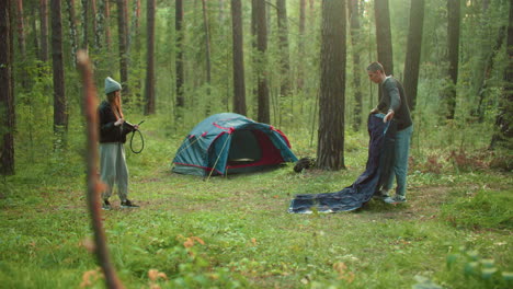 woman in knit beanie watches man unfold tent fabric on grassy forest clearing with pitched tent in background surrounded by trees as they prepare gear and equipment