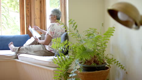 Glückliche-ältere-Frau-Mit-Gemischter-Abstammung-Auf-Der-Couch,-Die-Zu-Hause-Am-Fenster-Ein-Buch-Liest,-Zeitlupe