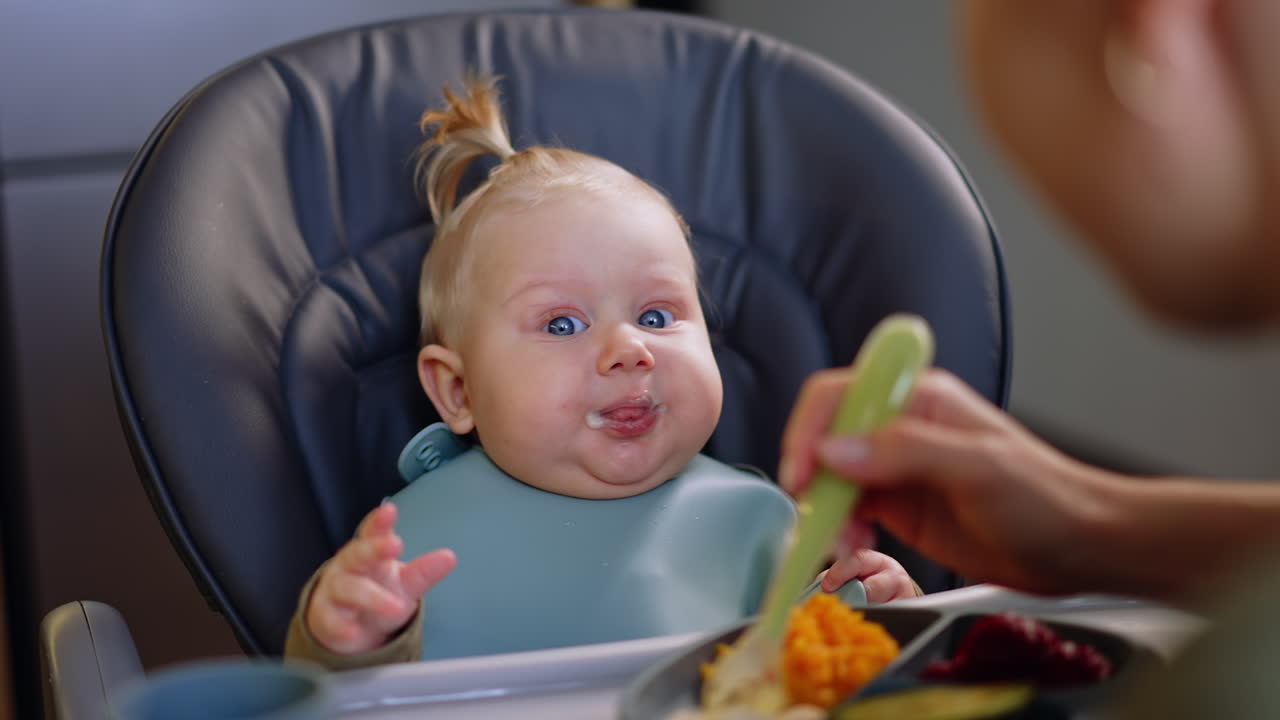 Feeding a cute little blond infant sitting in high chair. Unrecognized mom gives food on the spoon to her son.
