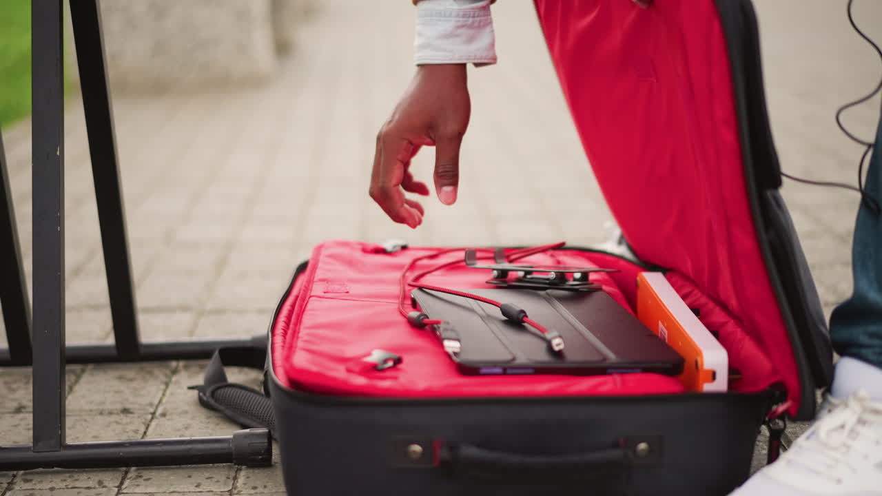 Gademusiker åbner rød kuffert med udstyr, hænder tilslutter controller til mixer, jeans og hvide sneakers synlige, fortov som baggrund, bypladsatmosfære, afslappet forberedelse til liveoptræden, kabler.