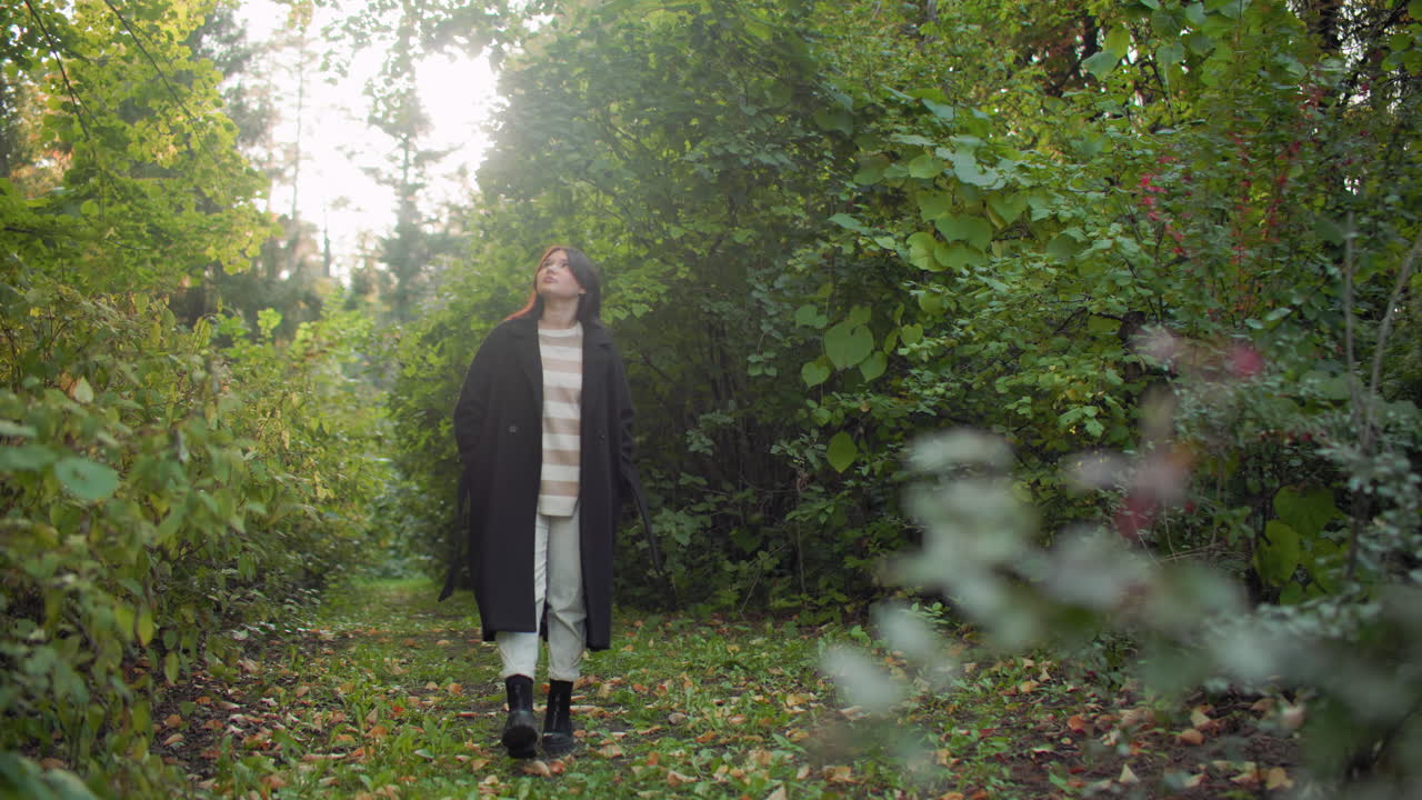 Fashionable girl strolling leisurely along green forest path, hands on jacket, gazing around with calm thoughtful expression surrounded by lush foliage under warm natural sunlight