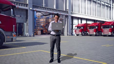 full body of an asian male professional worker standing with his laptop, outside of logistics distributions warehouse, working continuously