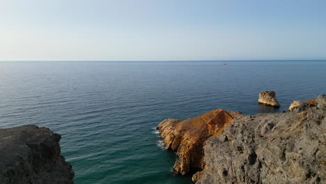 Una-Vista-Aérea-Panorámica-Captura-Un-Majestuoso-Acantilado,-Una-Escarpada-Playa-Rocosa-Y-Un-Carguero-Distante-En-El-Horizonte