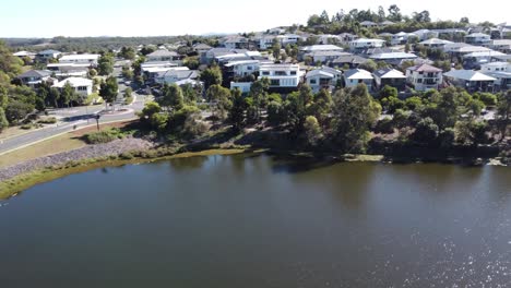 Drone-Ascendiendo-Sobre-Un-Lago-Artificial-Cerca-De-Un-Suburbio-Australiano-Con-Grandes-Casas-Familiares-Y-Una-Carretera-Con-Una-Rotonda