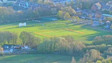 aerial static shot of a cricket match in progress on a bright green pitch near manvers lake, rotherham, with long shadows cast across the field and surrounding suburban buildings under the spring sun