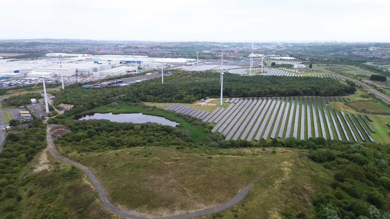 Cinematic aerial footage showing a large solar panel array and wind turbines integrated into a green landscape next to a modern industrial facility. Perfect for sustainability and clean energy concept