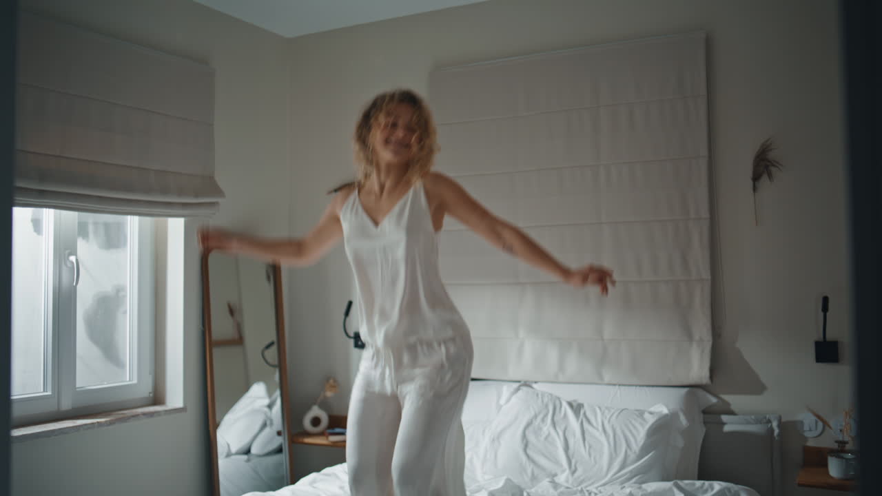 Donna spensierata che salta sul letto con i vestiti. Ragazza riccia felice che si allunga al mattino.