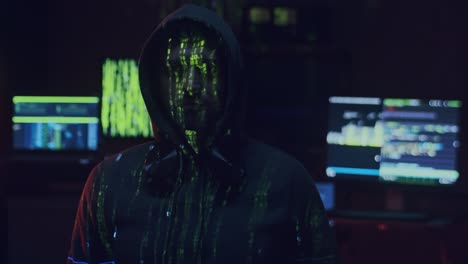 portrait shot of the young male software developer or hacker in a hood looking straight to the camera while running projected code numbers and characters on him in the dark room with computers and devices