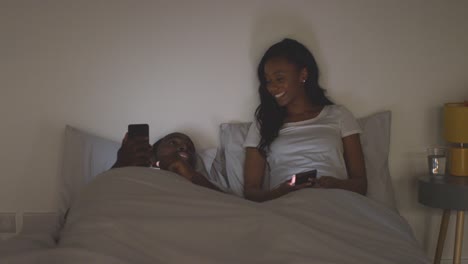 Junges-Paar-Entspannt-Sich-Nachts-Zu-Hause-Im-Bett-Und-Schaut-Gemeinsam-Auf-Mobiltelefone