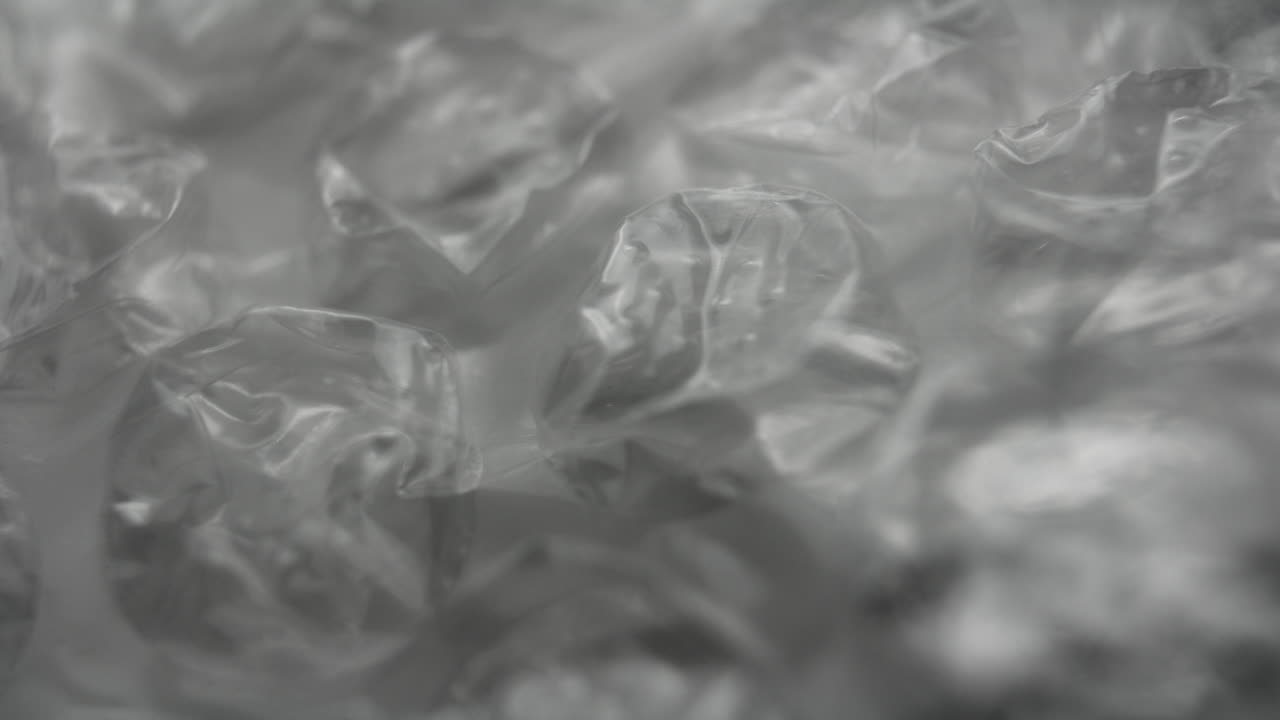 Primo piano astratto di pluriball trasparente con movimento lento della camera da sinistra a destra. Sfondo monocromatico minimal con texture di plastica, messa a fuoco morbida e riflessi di luce tenui.