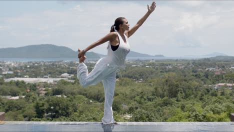 Barefooted-woman-in-standing-bow-pose-near-swimming-pool-against-green-forested-islands