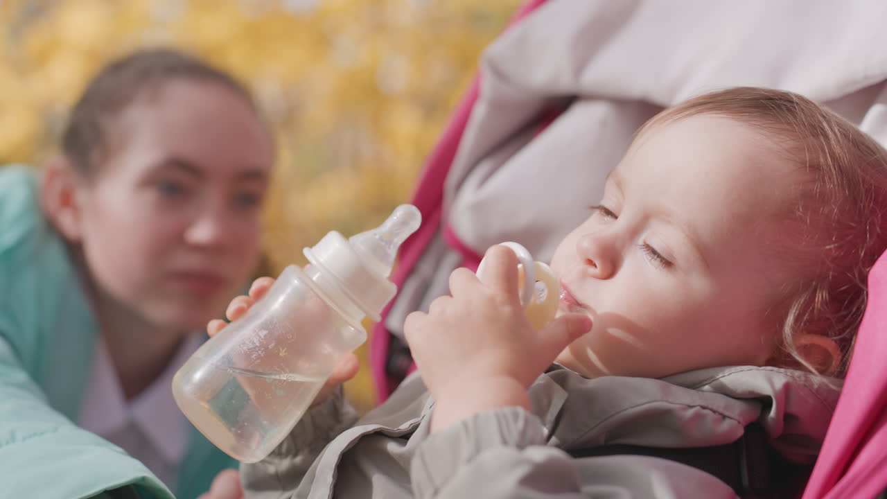 Bambino che cerca il biberon nel passeggino, madre bianca che osserva nelle vicinanze, ambientazione in un parco autunnale con foglie dorate, tentativo giocoso di auto-alimentarsi, scambio di ciuccio e biberon, cura familiare di supporto.