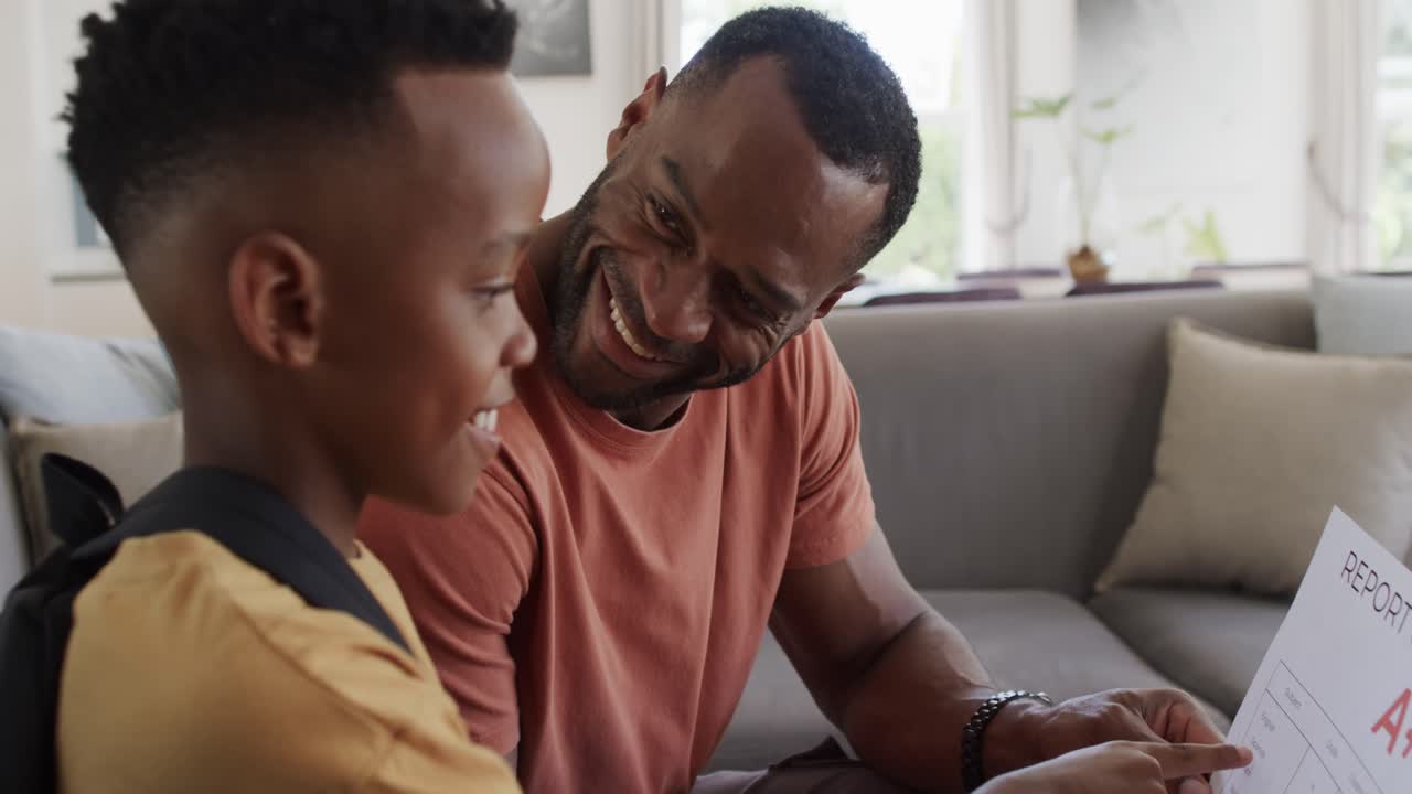 Father and son holding report card showing uppercase A+, pointing, high-fiving on living room couch