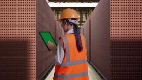 back view of asian female engineer with safety helmet working on a green screen tablet and looking around with red brick packed in stacks are stored