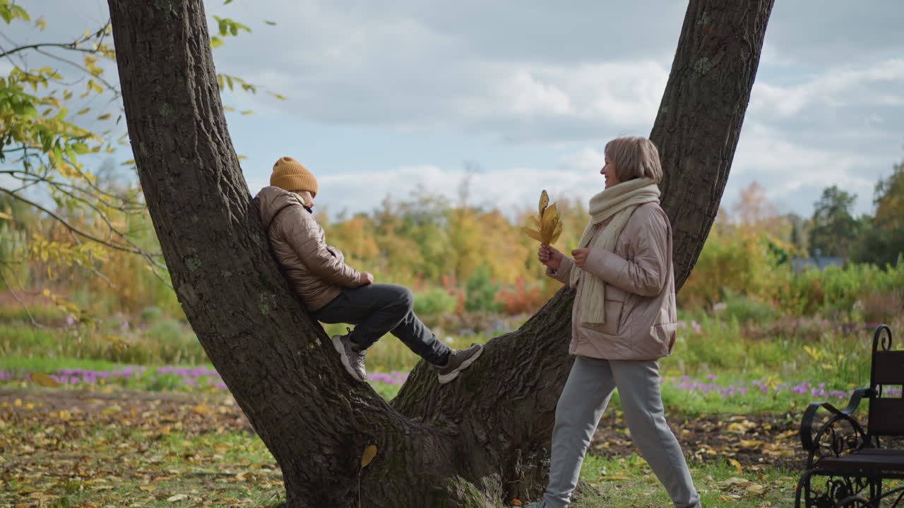 loving mother walks up to daughter sitting on tree in autumn park handing yellow leaf and kissing daughter on cheek creating tender family moment amid vibrant fall foliage and serene nature backdrop