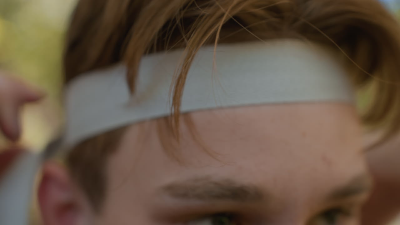 Close up view of tourist forehead and eyebrow while hands tie white headband, auburn hair strands framing skin, blurred woodland background and soft sunlight highlighting texture and focused motion