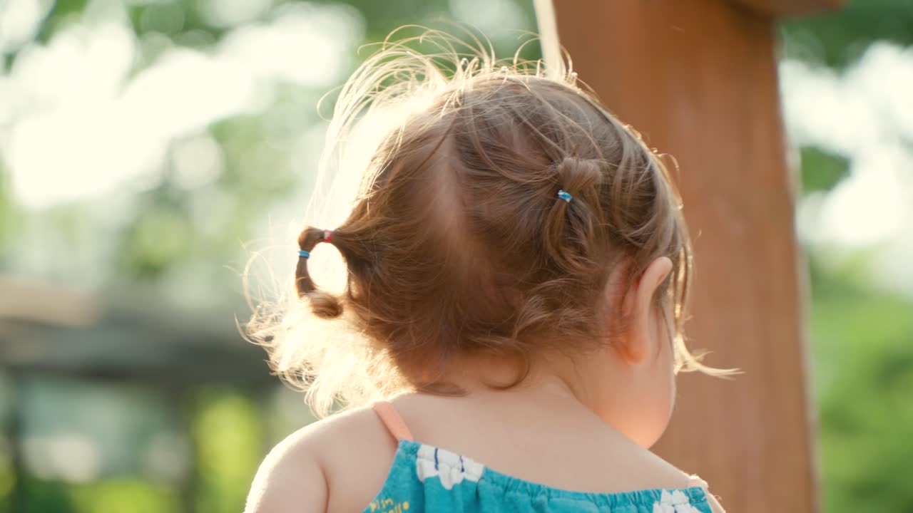niña pequeña sentada en un patio de recreo en la luz suave de la puesta de sol de verano y comiendo, girando la cabeza y mirando a la cámara - cámara lenta de enfoque superficial