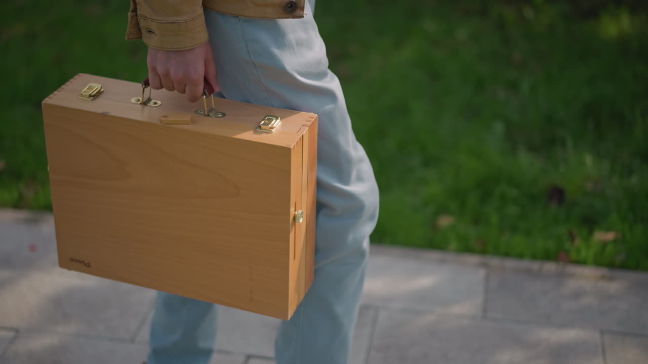 artista che trasporta una valigetta di legno sul marciapiede, jeans e scarpe da ginnastica visibili, scatola da schizzi con chiusura in ottone stretta in mano mentre cammina davanti al verde del parco, luce solare soffusa e ombra, scena di arte urbana di strada